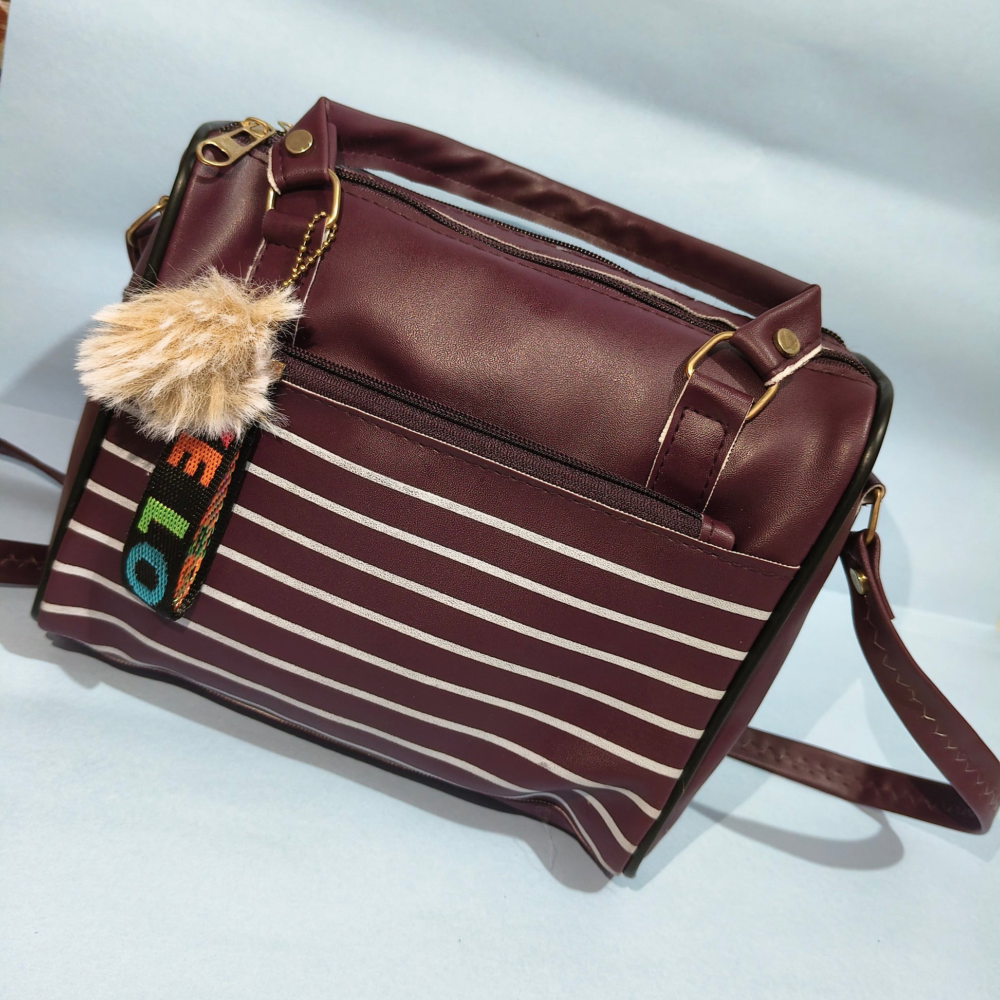 Brown handbag with striped pattern and pom-pom detail on a light gray background